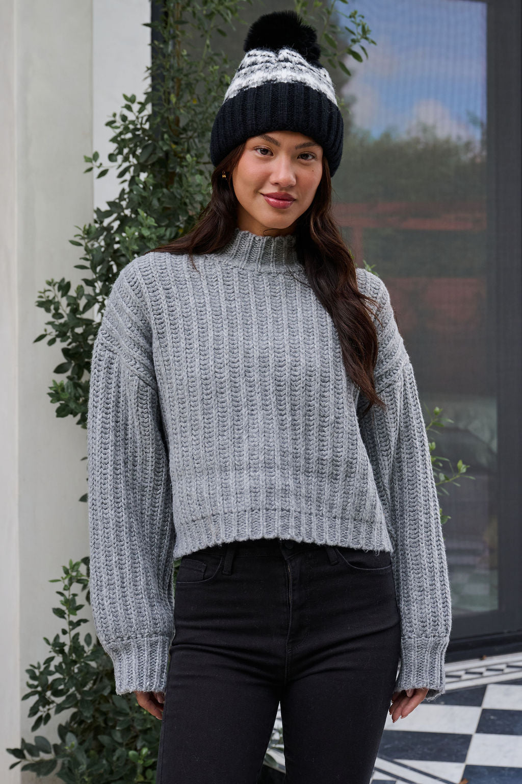 A woman wearing a Grey Wide Sleeve Ribbed Mock Neck Sweater, black pants, and a black winter hat stands outside, smiling.