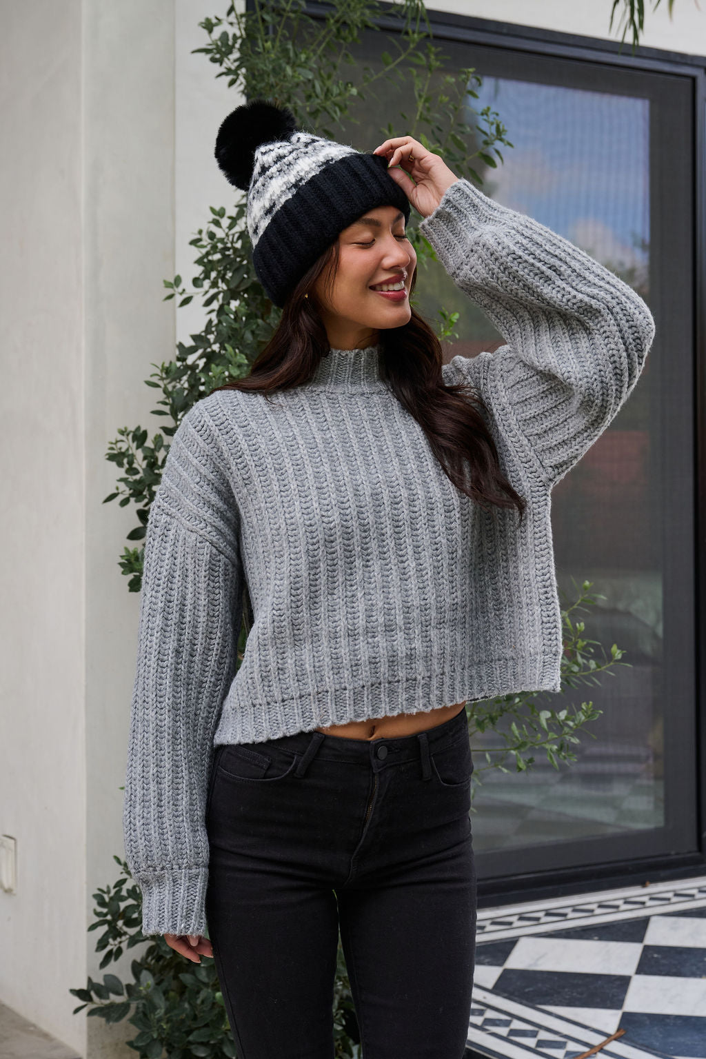 A woman in a Grey Wide Sleeve Ribbed Mock Neck Sweater and black beanie smiles outdoors, surrounded by greenery.