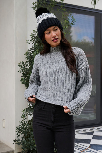 A woman in a Grey Wide Sleeve Ribbed Mock Neck Sweater, black pants, and winter hat poses outside a modern building.