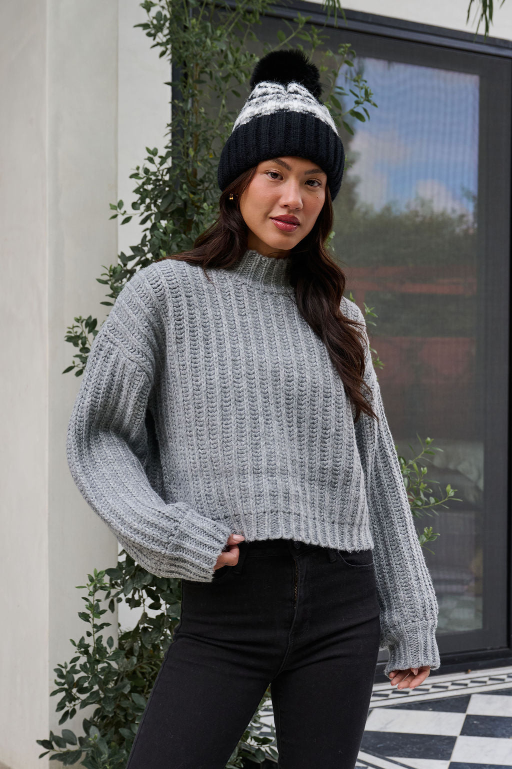 Woman in a Grey Wide Sleeve Ribbed Mock Neck Sweater, black pants, and beanie stands outdoors near greenery and a window.