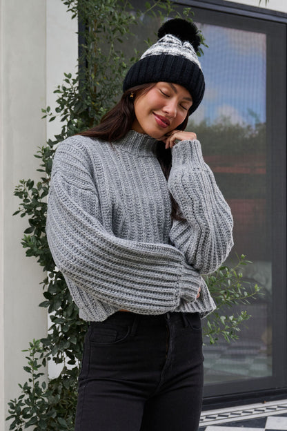 Woman in a Grey Wide Sleeve Ribbed Mock Neck Sweater and black beanie smiles outdoors in front of a plant.