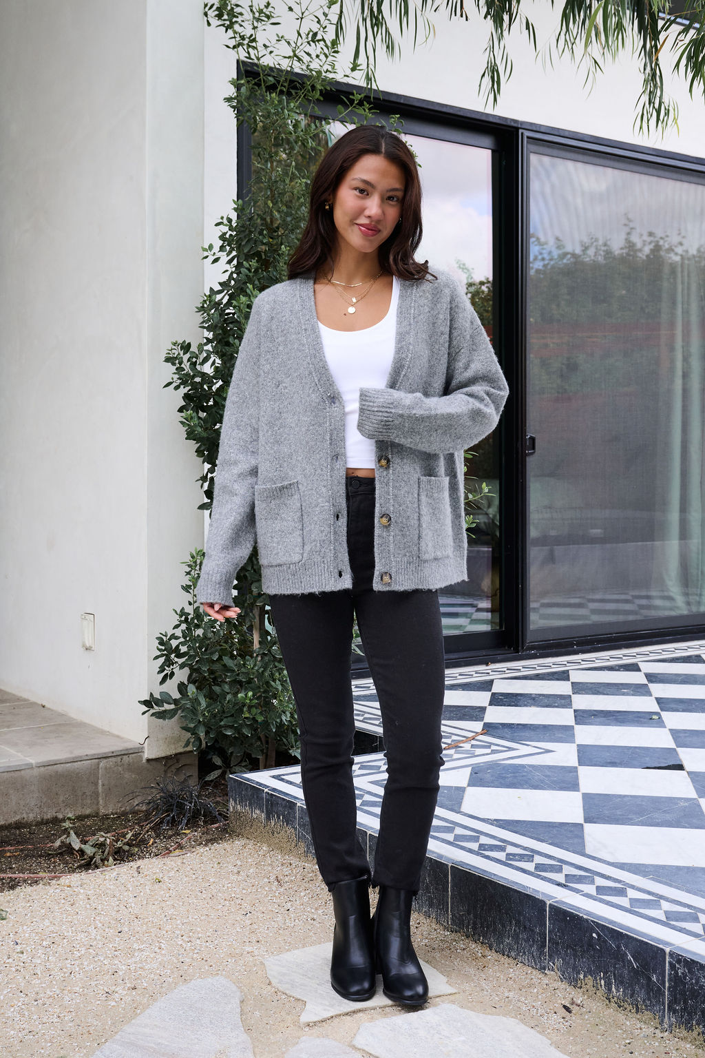 A woman stands on a tiled patio, smiling in the Cozy Grey Boyfriend Cardigan, white top, black pants, and black boots.