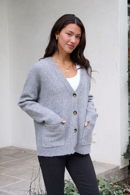 A woman stands outside in a Cozy Grey Boyfriend Cardigan, white top, and black pants with hands in cardigan pockets.