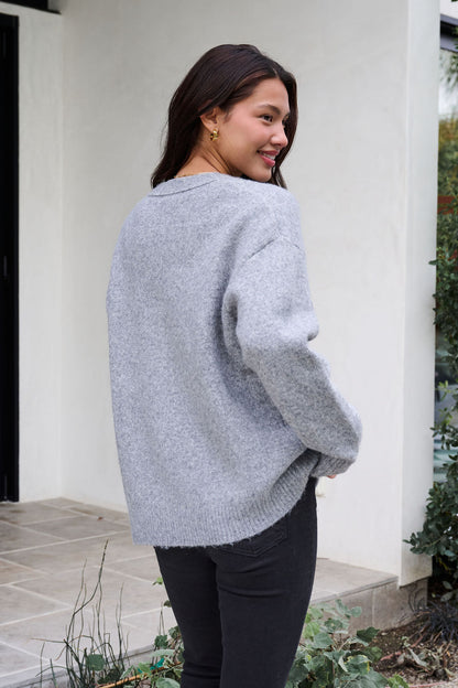 Woman with long dark hair smiles outdoors in a Cozy Grey Boyfriend Cardigan and black pants, turned slightly from the camera.