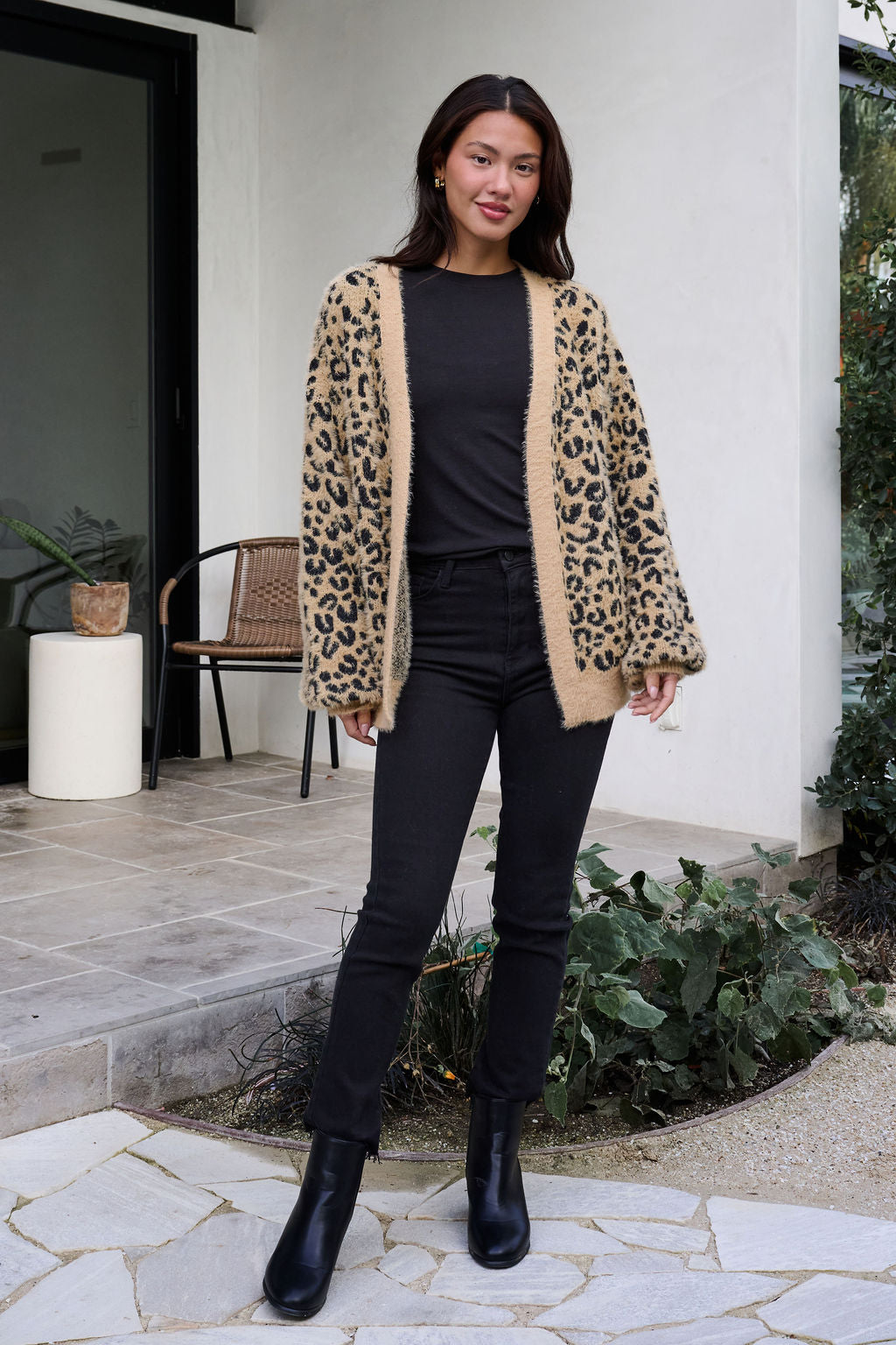 Woman stands outdoors in a Fuzzy Tan Leopard Open Front Cardigan, black top and pants, with plants and a chair behind.