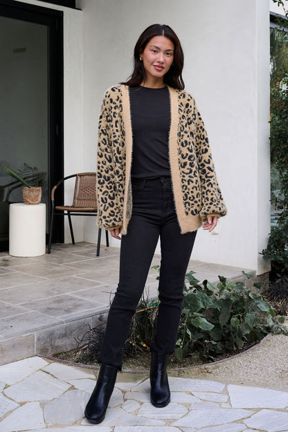 Woman stands outdoors in a Fuzzy Tan Leopard Open Front Cardigan, black top and pants, with plants and a chair behind.