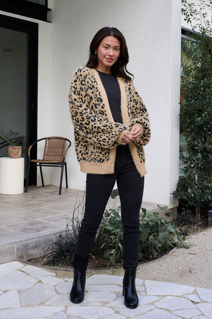 Woman outside in a Fuzzy Tan Leopard Open Front Cardigan, black top, jeans, and boots, near plants and a chair.
