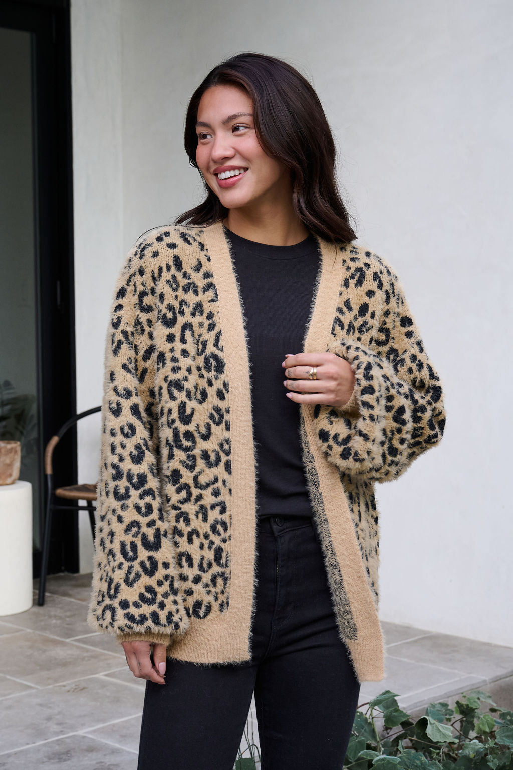 A woman smiles outdoors in a Fuzzy Tan Leopard Open Front Cardigan layered over a black shirt and pants.