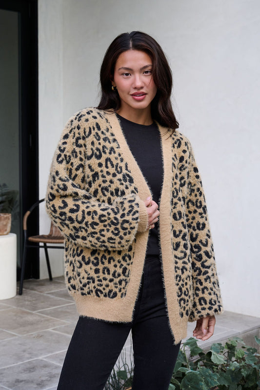 Woman outdoors wearing a Fuzzy Tan Leopard Open Front Cardigan over black, with plants and patio furniture in the background.
