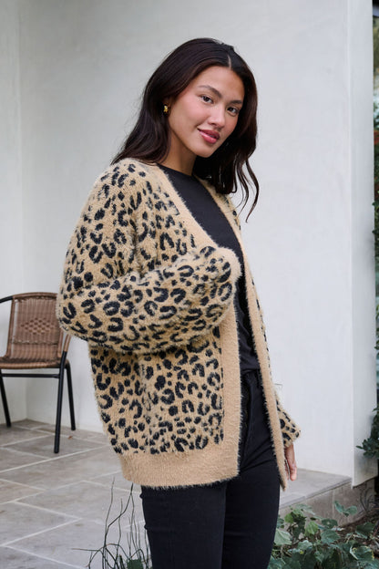 A woman wearing a Fuzzy Tan Leopard Open Front Cardigan and black pants stands outside by a chair, posing and smiling slightly.