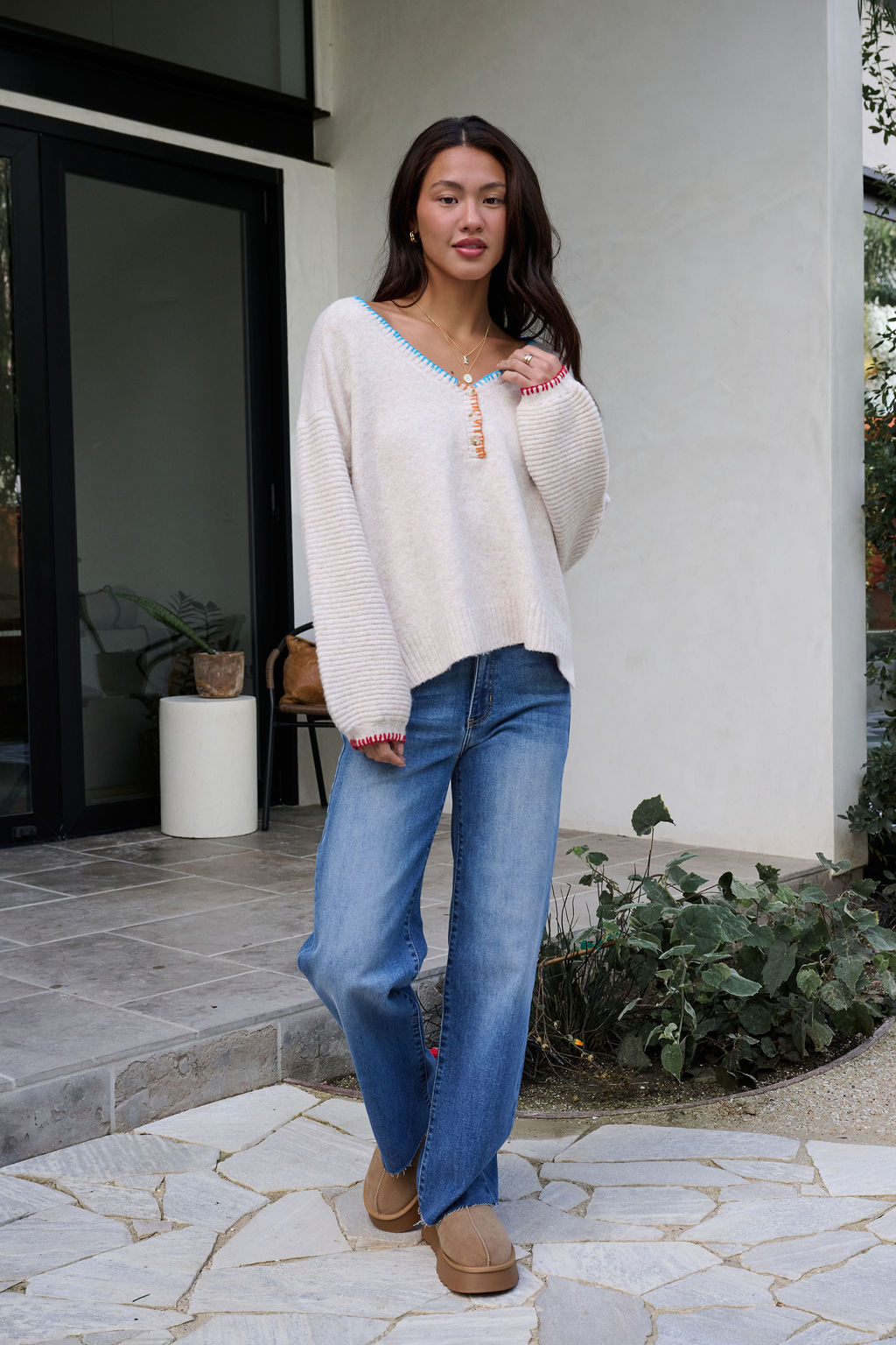 A woman in an Ivory Stitch Trim Button Up Sweater, blue jeans, and tan shoes stands outside a modern building with plants nearby.