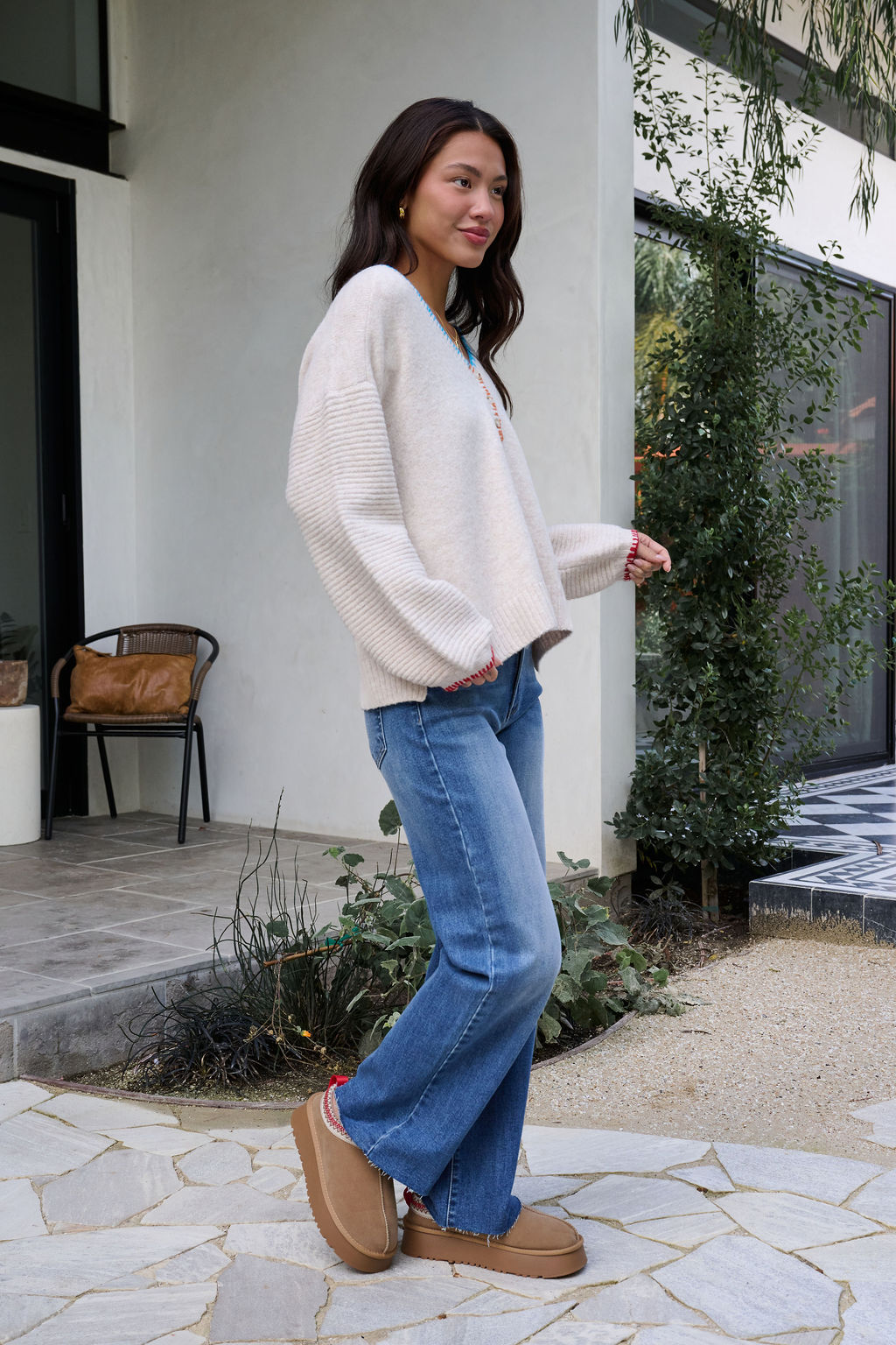 A woman in an Ivory Stitch Trim Button Up Sweater, blue jeans, and tan slippers smiles outdoors on a stone patio.