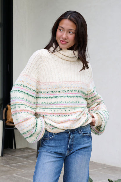 Smiling woman stands outside in blue jeans and a Cozy Cream Turtleneck Striped Sweater, with one hand in her pocket.