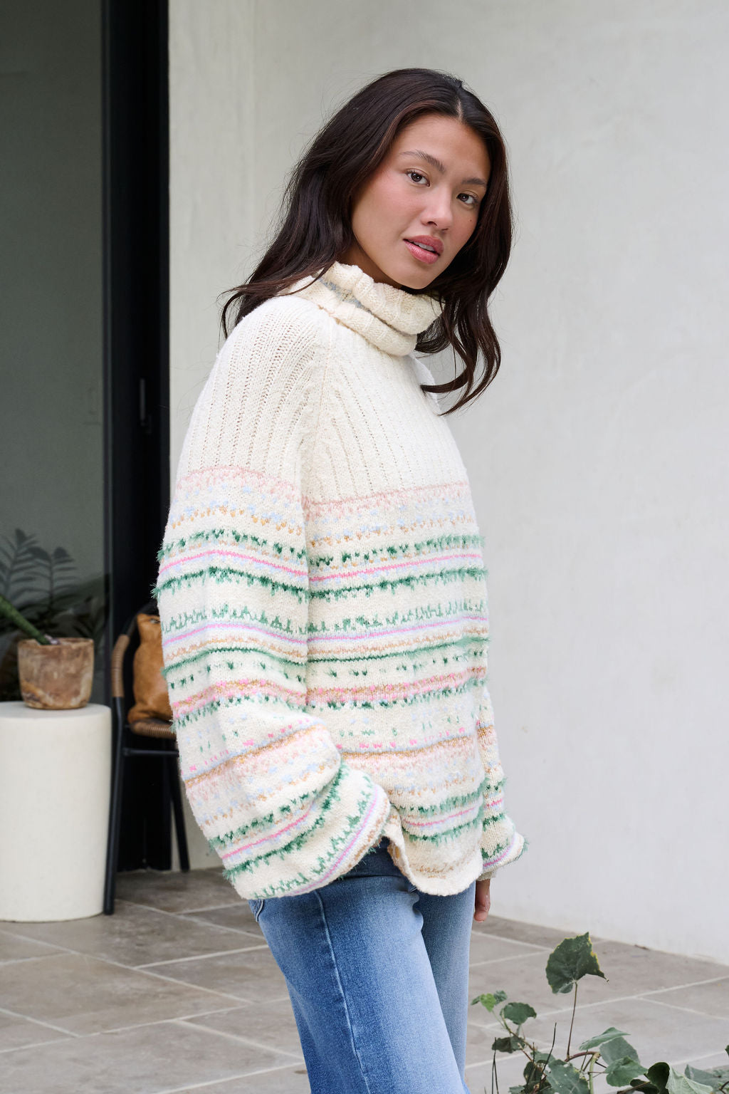 A woman in the Cozy Cream Turtleneck Striped Sweater stands on a tiled patio by plants, bringing festive charm to the scene.