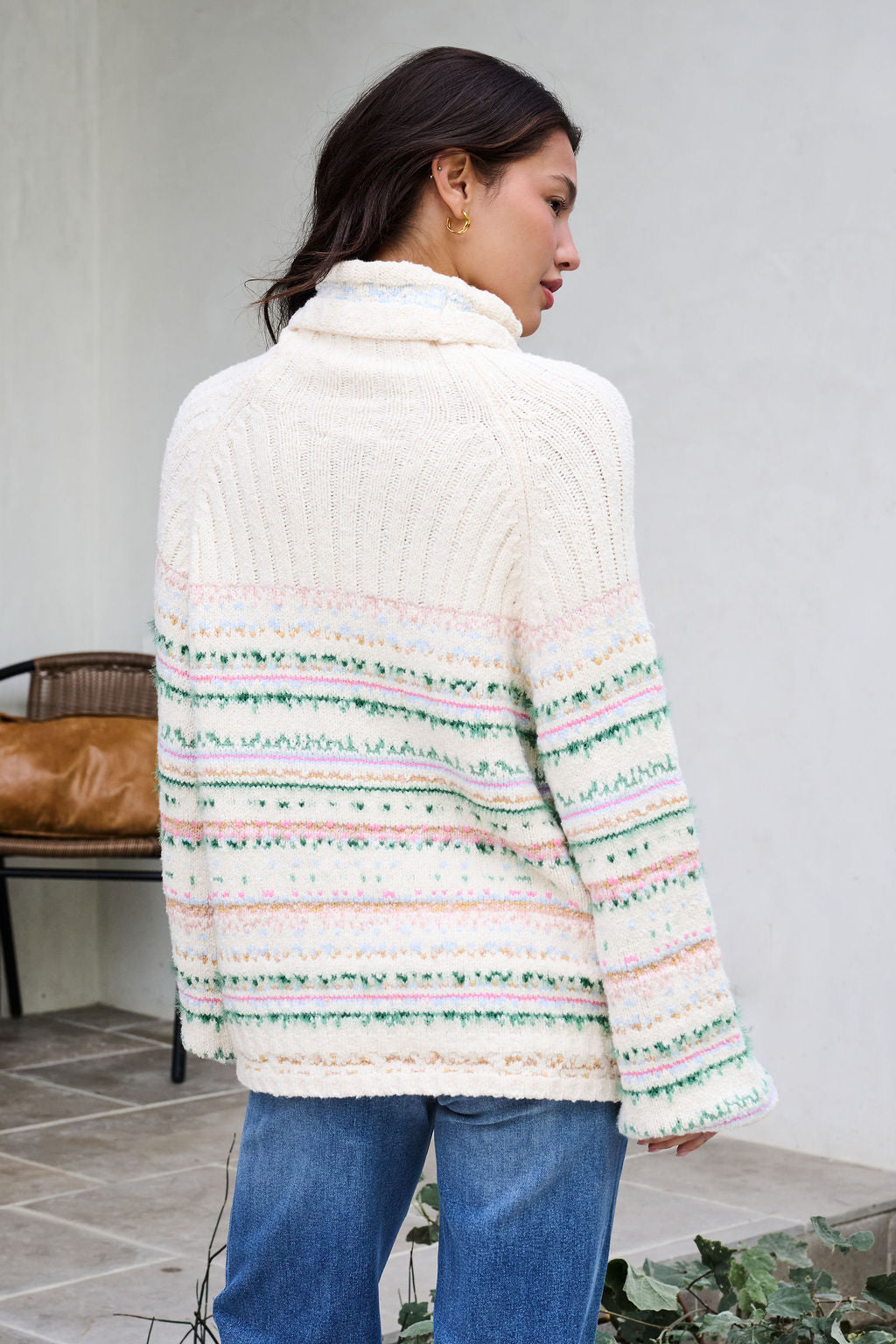 Woman with long dark hair wears the Cozy Cream Turtleneck Striped Sweater and blue jeans outdoors, facing away for holiday charm.