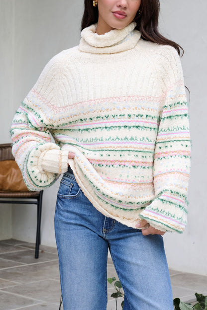 A woman in a Cozy Cream Turtleneck Striped Sweater and blue jeans stands by a chair indoors, showcasing festive holiday style.