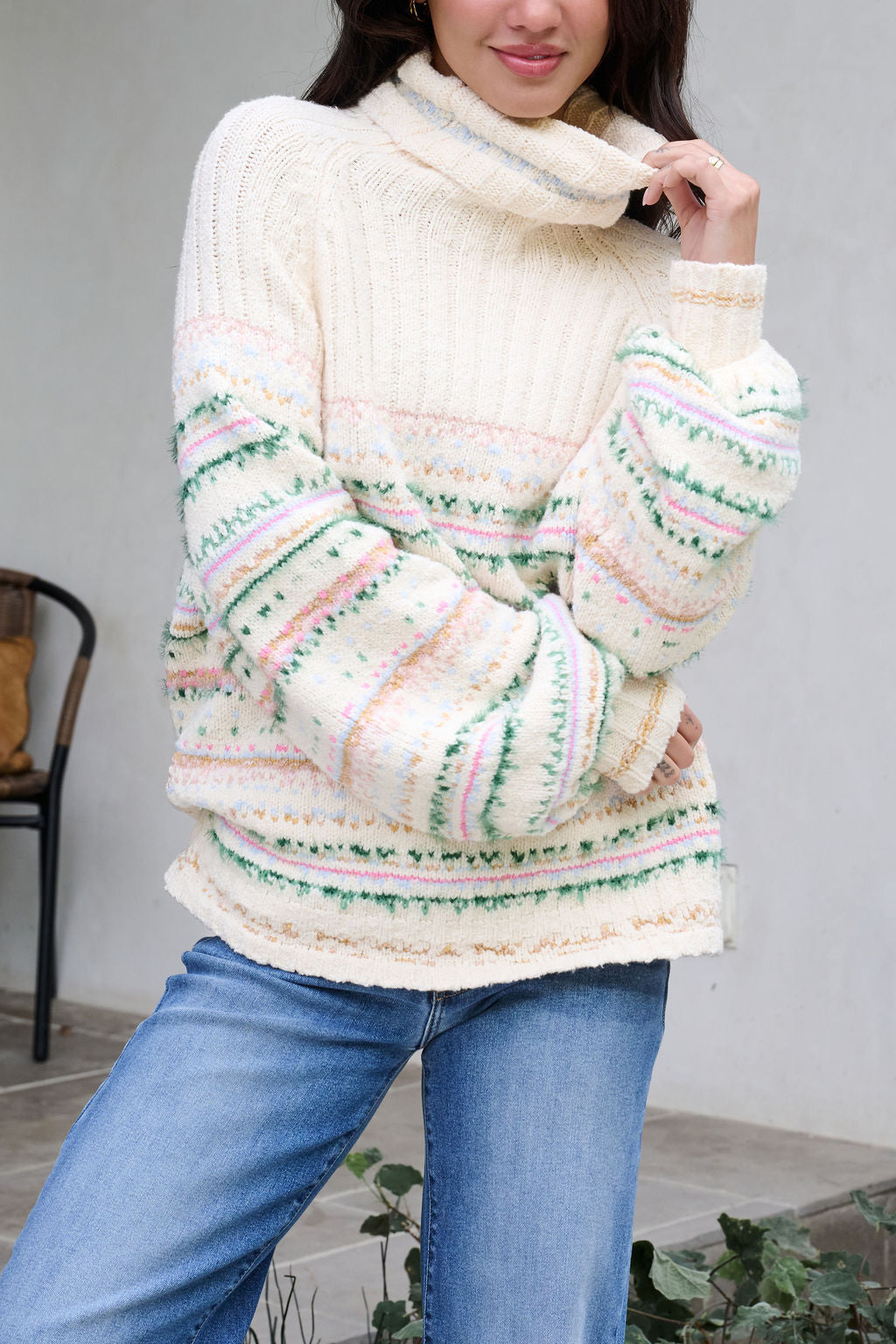 Woman in a Cozy Cream Turtleneck Striped Sweater with pastel stripes and blue jeans, radiating holiday spirit indoors by a chair.