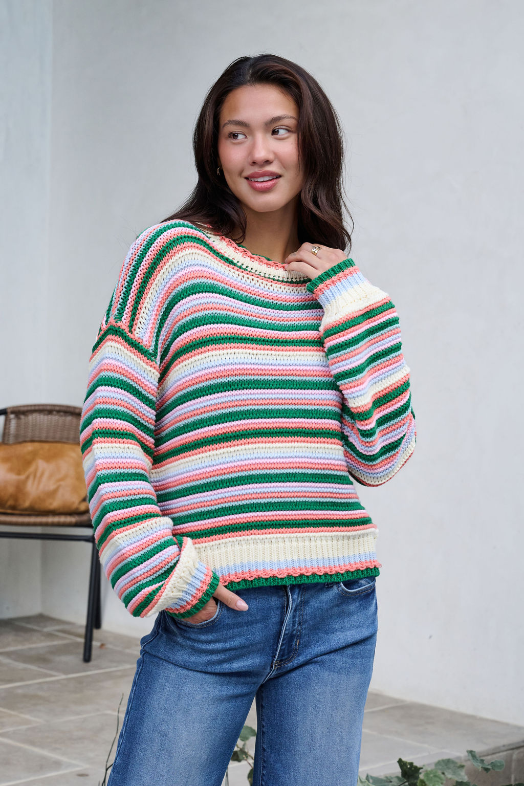 A woman in an Ivory Multi Striped Crewneck Sweater and blue jeans stands outdoors, smiling with one hand in her pocket.