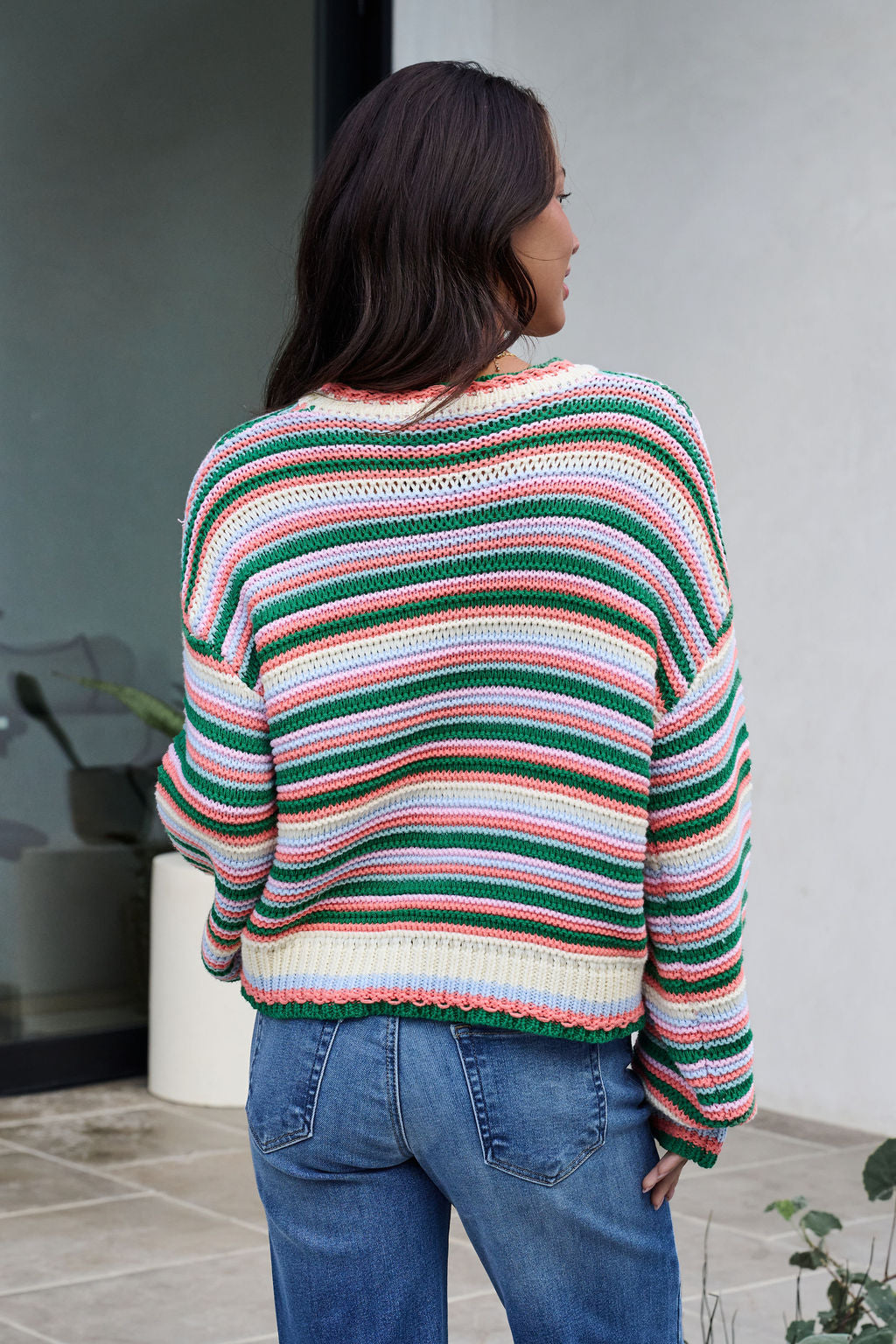 A woman with long dark hair, seen from behind, wears an Ivory Multi Striped Crewneck Sweater and blue jeans outdoors.