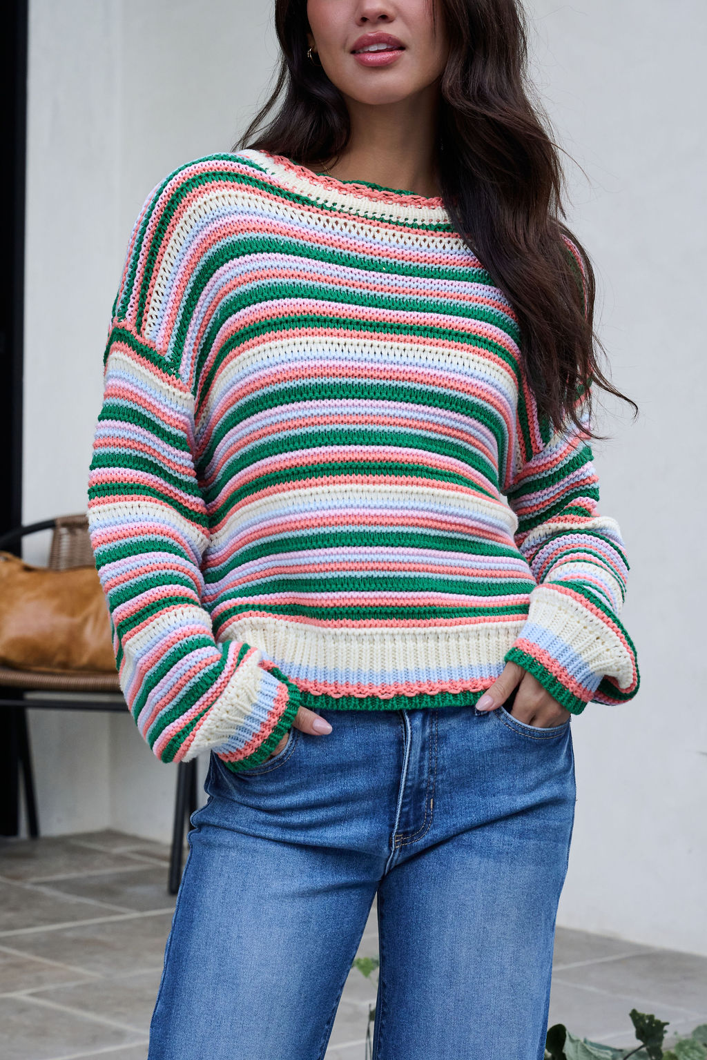 Woman wears an Ivory Multi Striped Crewneck Sweater and blue jeans outdoors, hands in pockets near a chair and bag.