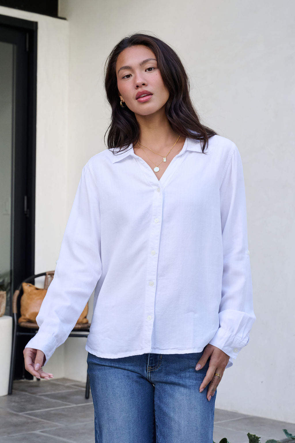 A woman in a Thread and Supply White Button Down Presley Top and blue jeans stands outside by a window, looking at the camera.