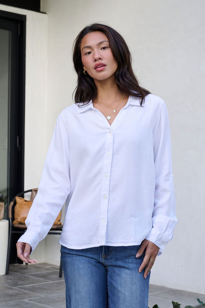 A woman in a Thread and Supply White Button Down Presley Top and blue jeans stands outside by a window, looking at the camera.