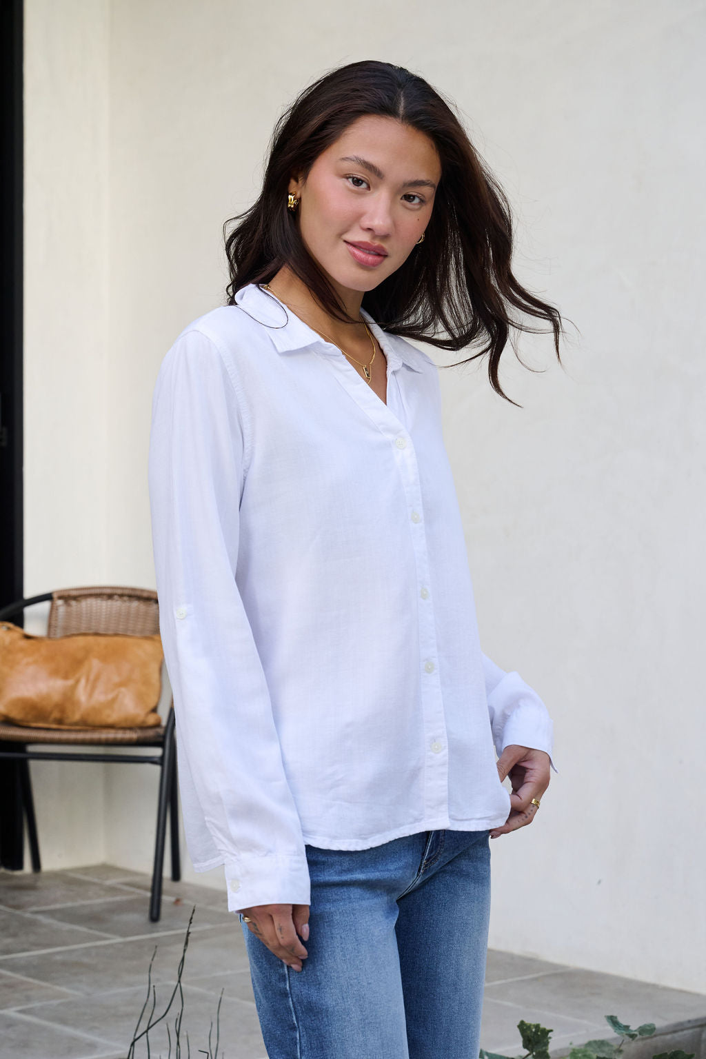 A woman in a Thread and Supply White Button Down Presley Top stands outside in blue jeans, smiling at the camera.