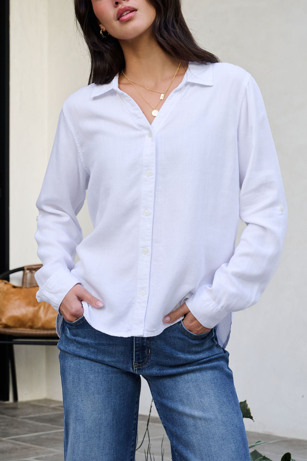 Woman in a Thread and Supply White Button Down Presley Top and blue jeans stands with hands in pockets, bench and bag behind her.