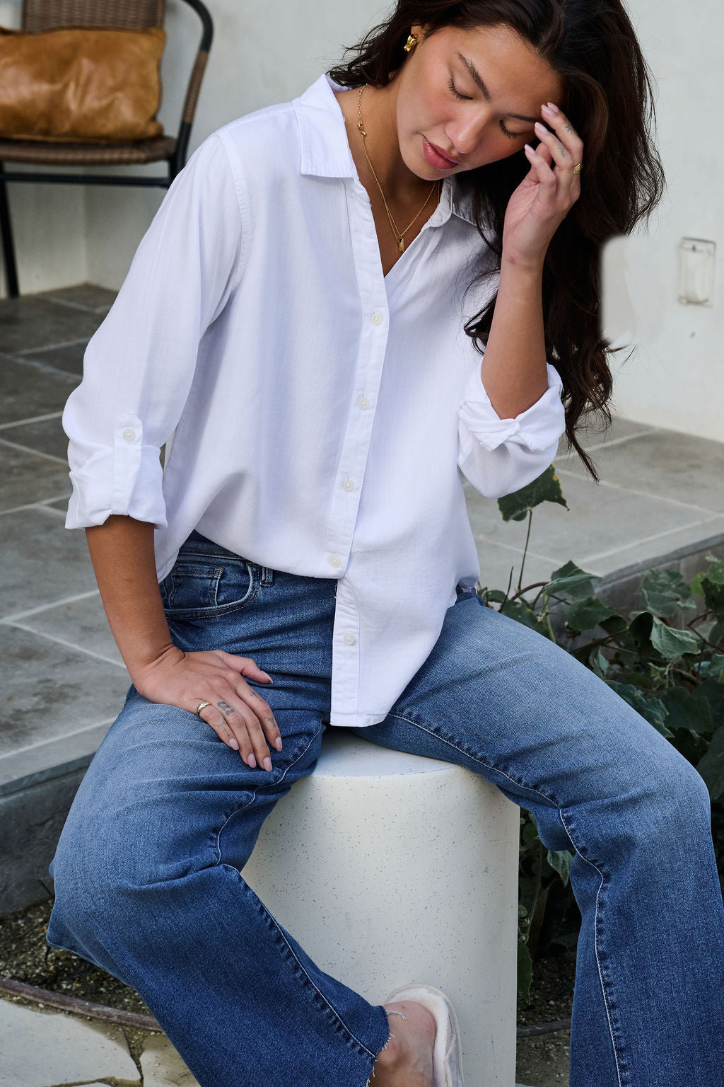 A woman in a Thread and Supply White Button Down Presley Top and blue jeans sits on a white stool outdoors, touching her hair.