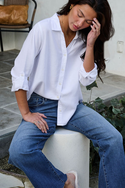 A woman in a Thread and Supply White Button Down Presley Top and blue jeans sits on a white stool outdoors, touching her hair.