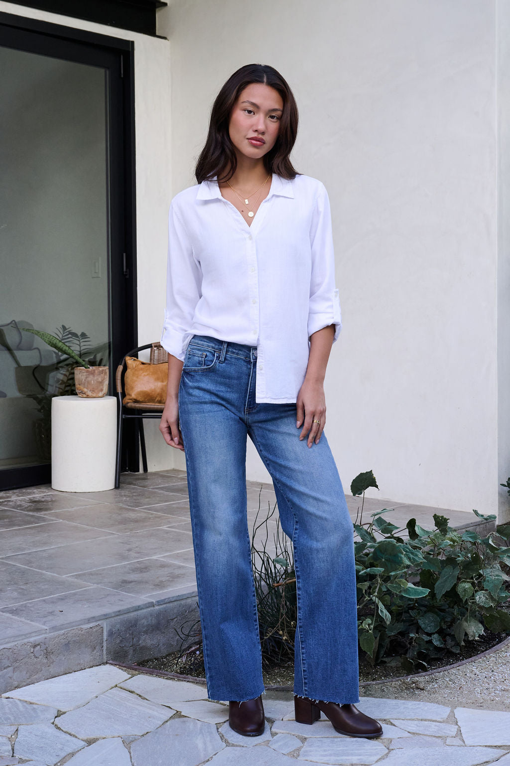 Woman stands on a stone patio in the Thread and Supply White Button Down Presley Top, blue jeans, and brown boots with plants behind her.