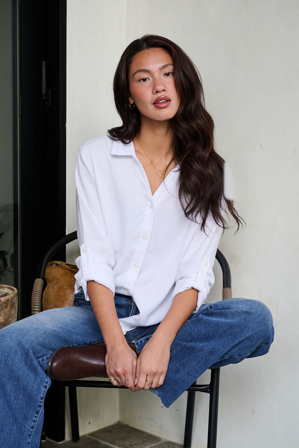 Woman in Thread and Supply Presley White Button Down sits casually on a chair in blue jeans against a light wall.