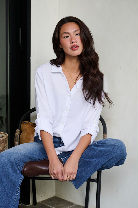 Woman in Thread and Supply Presley White Button Down sits casually on a chair in blue jeans against a light wall.