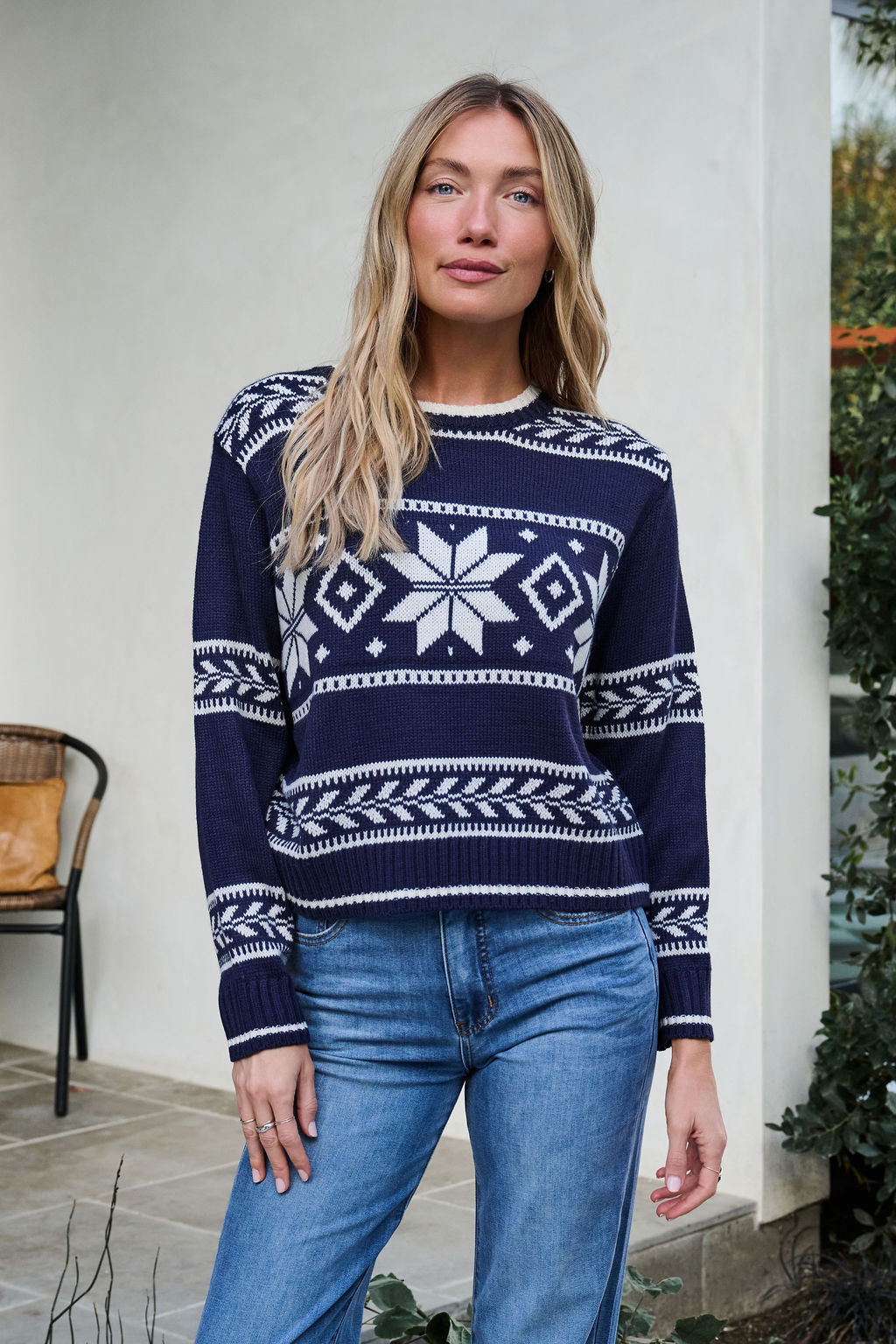 Woman with long blonde hair wears a Navy Ribbed Winter Snowflake Sweater and blue jeans, standing outdoors near greenery.