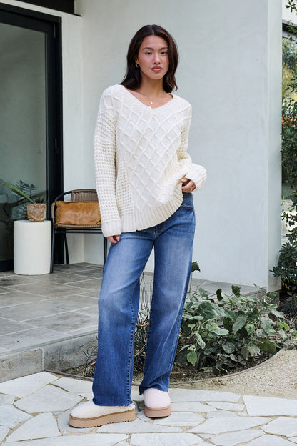 A woman in an Ivory Textured Cable Knit Sweater, blue jeans, and beige slippers stands outside a modern house near plants.