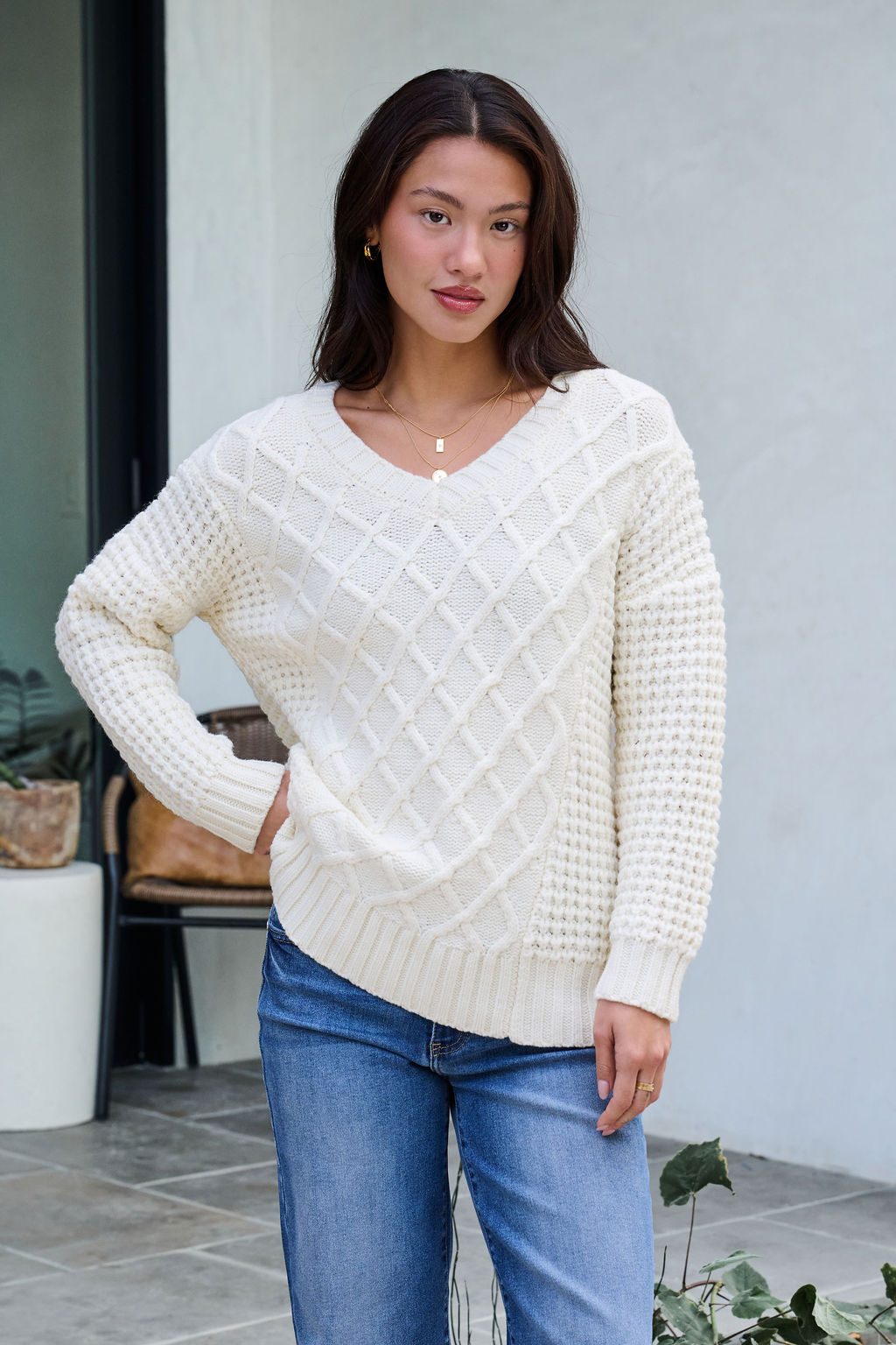 Woman with long brown hair wears an Ivory Textured Cable Knit Sweater and blue jeans, standing outdoors, hand on hip.