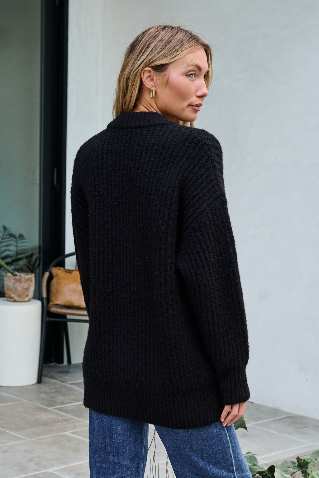 A woman with blonde hair wears a Black Heavy Knit Button Down Cardigan and blue jeans as she stands outside, looking over her shoulder.