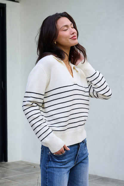 Woman outside in an Ivory and Black Stripe Ribbed Sweater and jeans, smiling with eyes closed, touching her hair.