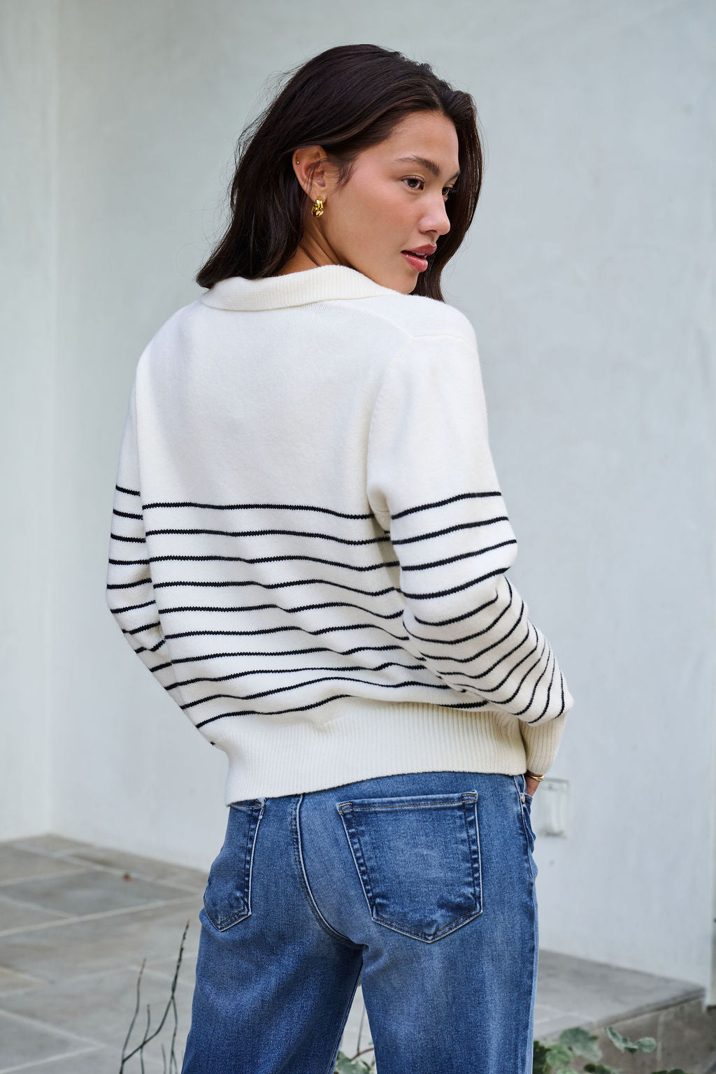 A woman wearing the Ivory and Black Stripe Ribbed Sweater with blue jeans stands outside, looking over her shoulder.