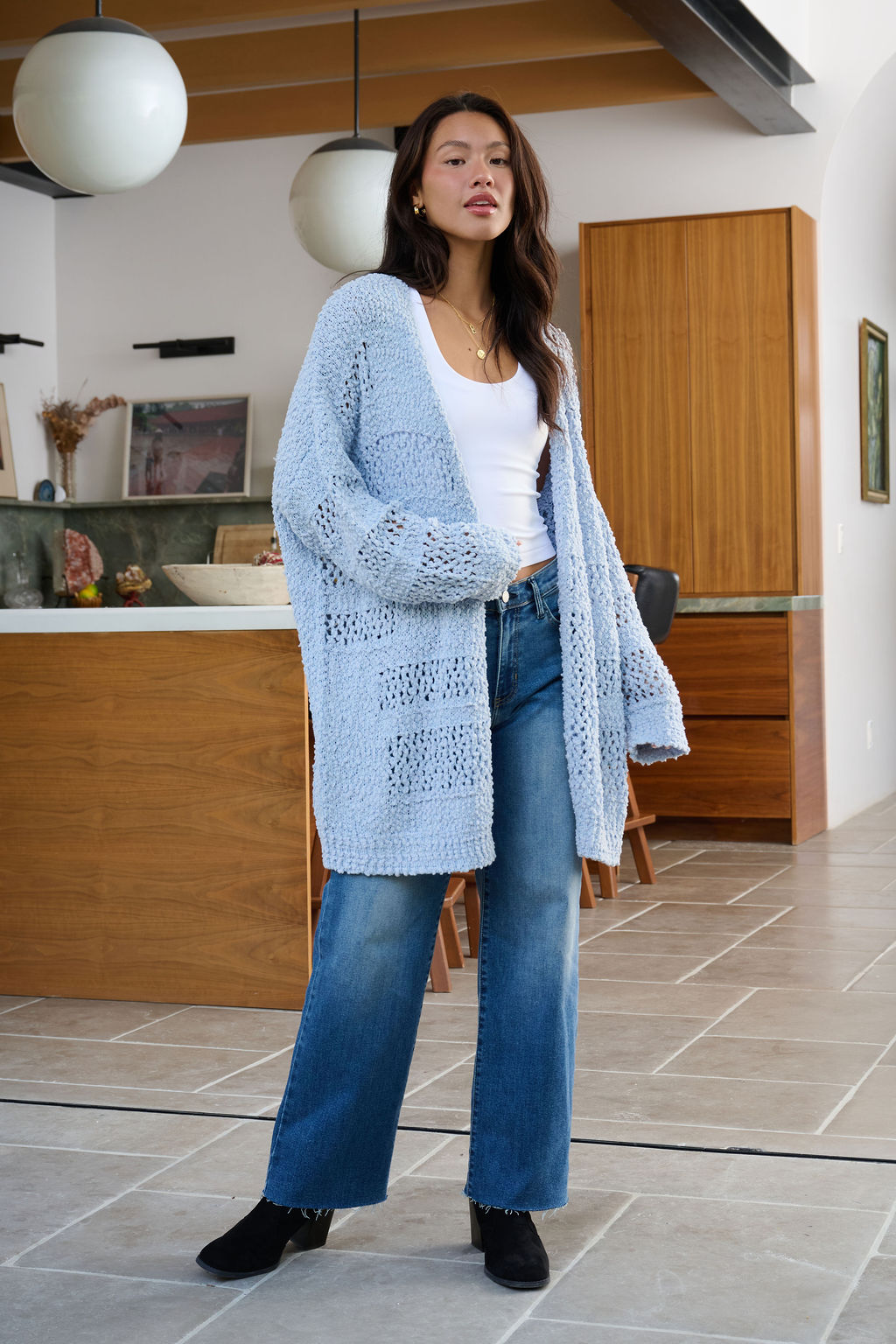 A woman in a white top, blue jeans, and a Blue Open Front Popcorn Knit Cardigan stands in a modern living room with wooden accents.