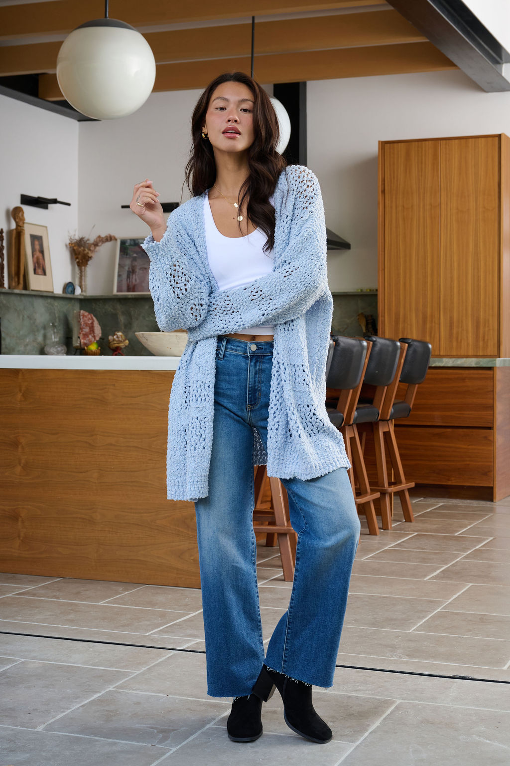 Woman stands in a modern kitchen wearing a Blue Open Front Popcorn Knit Cardigan, white top, blue jeans, and black shoes.