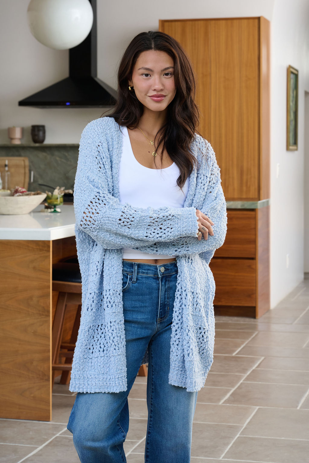 A woman in a Blue Open Front Popcorn Knit Cardigan and jeans smiles with crossed arms in a modern kitchen.
