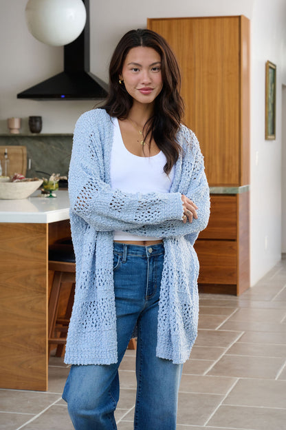 A woman in a Blue Open Front Popcorn Knit Cardigan and jeans smiles with crossed arms in a modern kitchen.