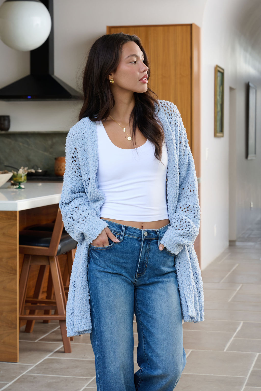 Woman in a white tank top, blue jeans, and a Blue Open Front Popcorn Knit Cardigan stands in a modern kitchen, looking to the side.
