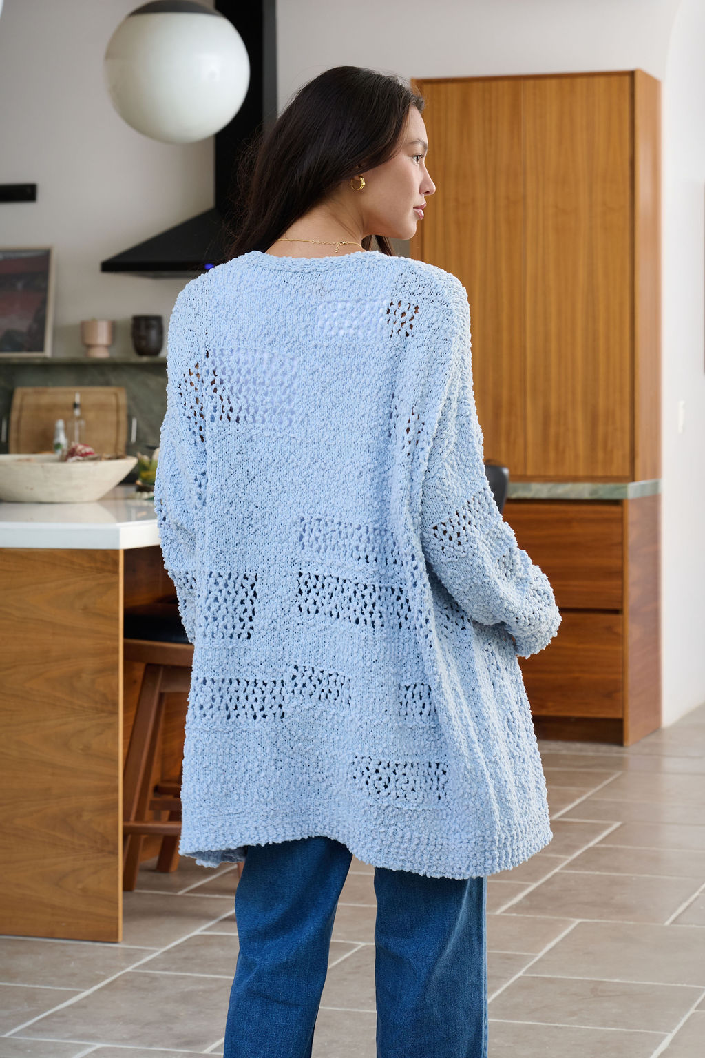 A woman with long dark hair wears a Blue Open Front Popcorn Knit Cardigan and blue jeans in a modern kitchen.