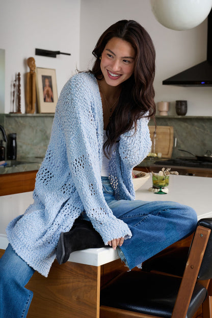 A woman in a Blue Open Front Popcorn Knit Cardigan and jeans sits smiling on a kitchen counter, legs crossed.