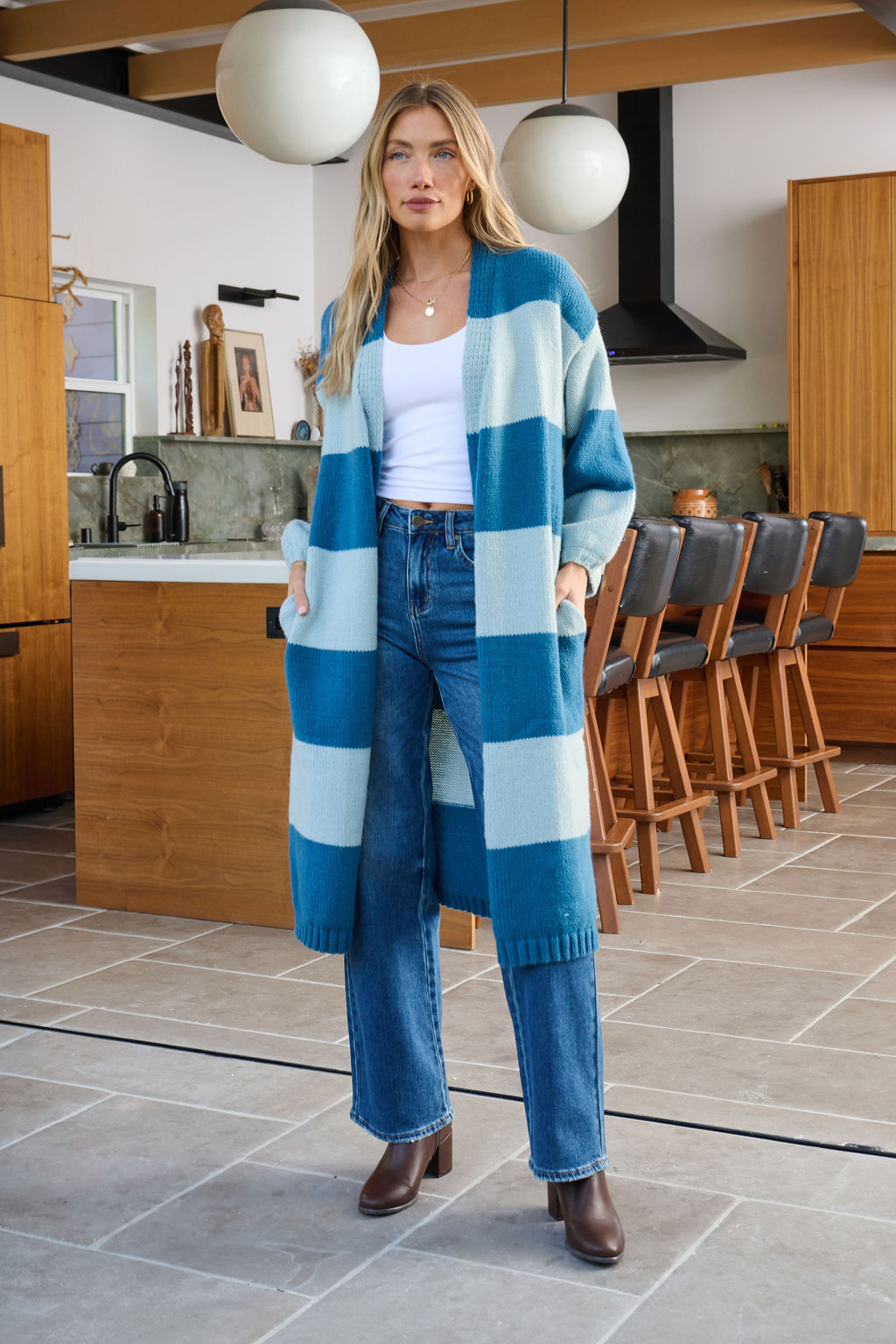 A woman in a Blue Striped Open Front Duster Cardigan, jeans, and boots stands in a modern kitchen with wooden cabinets.