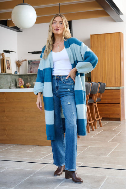 A woman in a Blue Striped Open Front Duster Cardigan, white top, and jeans stands in a modern kitchen with wooden cabinets.