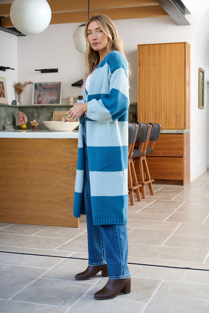 A woman in a Blue Striped Open Front Duster Cardigan, jeans, and brown boots stands in a modern kitchen with wooden cabinets.
