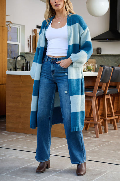 A woman in a Blue Striped Open Front Duster Cardigan and jeans stands in a modern kitchen—ideal for winter style.